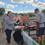 Hotdogs on a BBQ grill outside while four people stand around it. Nina Yoon and Jeannine Schellenberg are holding hotdogs in their hands and smiling at the camera.