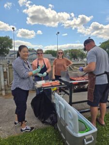 Hotdogs on a BBQ grill outside while four people stand around it. Nina Yoon and Jeannine Schellenberg are holding hotdogs in their hands and smiling at the camera.