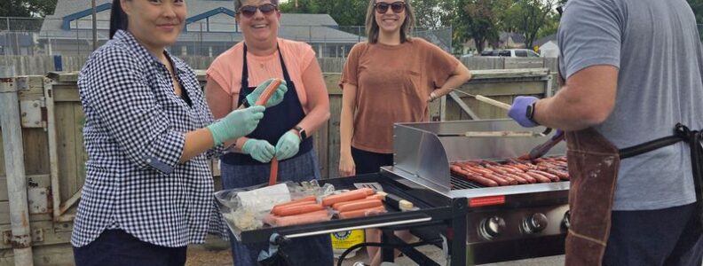 Hotdogs on a BBQ grill outside while four people stand around it. Nina Yoon and Jeannine Schellenberg are holding hotdogs in their hands and smiling at the camera.