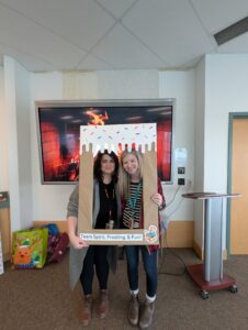 Meaghan & Amanda standing inside a carboard picture frame made to look covered in frosting. It says "Team Spirit, Frosting, & Fun!"