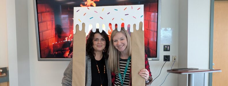 Meaghan & Amanda standing inside a carboard picture frame made to look covered in frosting. It says "Team Spirit, Frosting, & Fun!"